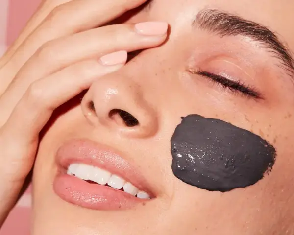 Una mujer sonríe con una mascarilla facial de arcilla negra en la mejilla. Después de una mascarilla facial, hay que ponerse crema para hidratar la piel.