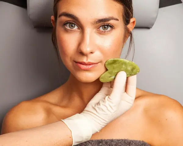 Una mano enguantada aplica una piedra de jade en la mejilla de una mujer, mostrando los beneficios del masaje facial para una piel radiante.