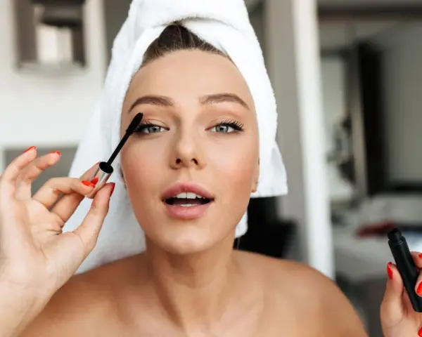 Mujer con toalla en la cabeza aplicándose máscara de pestañas como parte de su rutina maquillaje.