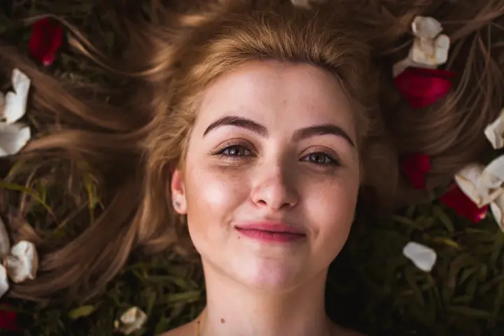Rostro de mujer joven con pecas y cejas naturales sin depilar, rodeada de pétalos de rosa y hojas.