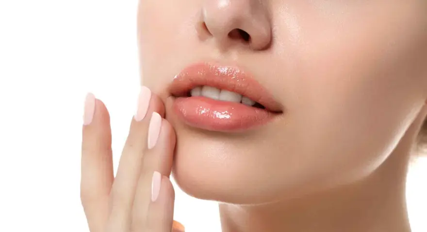 Labios rosados y brillantes, con una mano de uñas pálidas tocándolos suavemente. Un ejemplo perfecto de como hidratar los labios para un look jugoso.