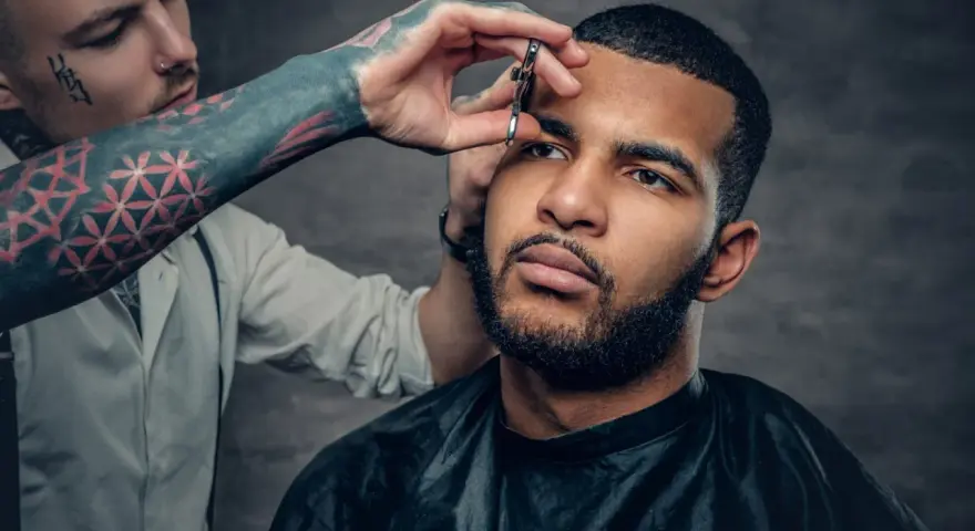 Barbero con tatuajes en el brazo recorta el **buzz cut corto** de un hombre con barba.