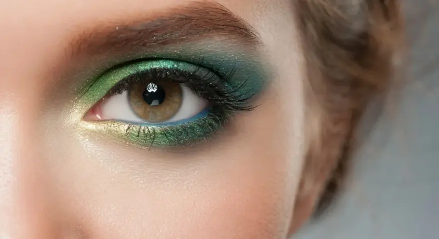 Ojo con maquillaje verde esmeralda y dorado, delineado azul y cejas verdes.