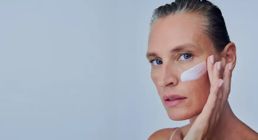 Mujer se aplica crema en la mejilla, como si fuera a quitar autobronceador. Suave toque para una piel radiante.