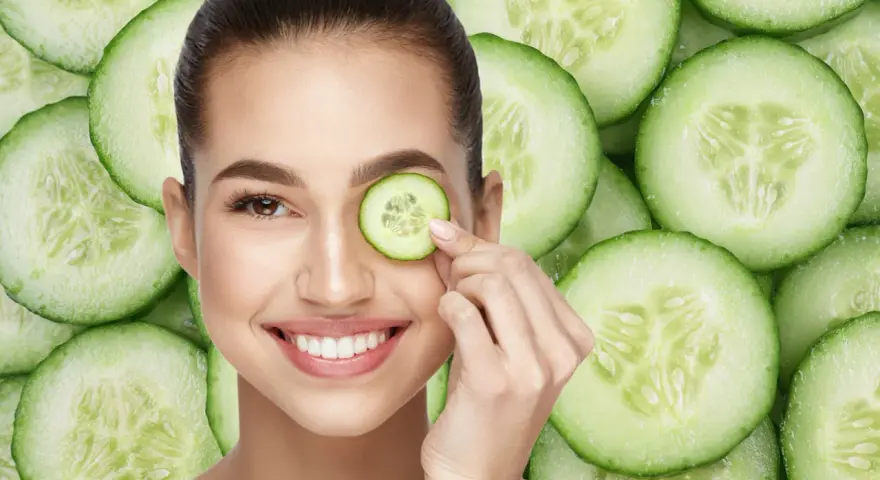 Una mujer sonríe mientras se coloca una rodaja de pepino en el ojo, rodeada de más rodajas de pepino.