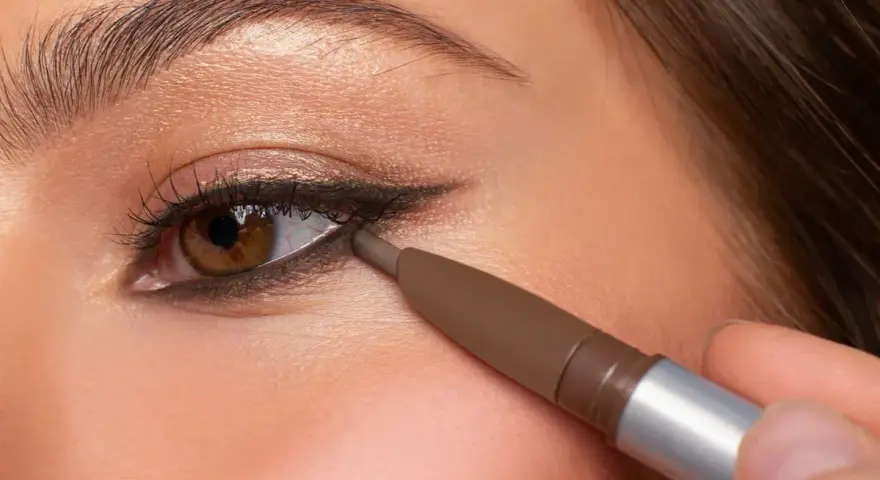 Ojo marrón aplicando delineador en la línea de agua inferior, creando un look de **reverse eyeliner** con sombra dorada en el párpado.