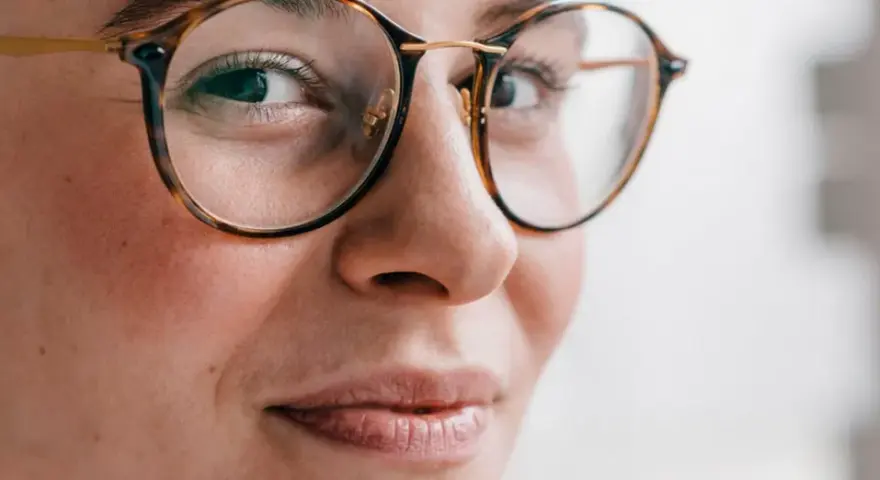 Primer plano de una mujer con gafas de montura de carey, sus ojos hundidos miran hacia un lado con una leve sonrisa.