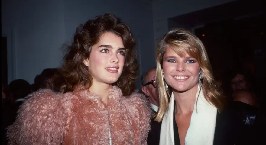 Dos mujeres con peinados icónicos de los 80, una con cabello castaño voluminoso y otra con rubio liso, posan sonrientes.