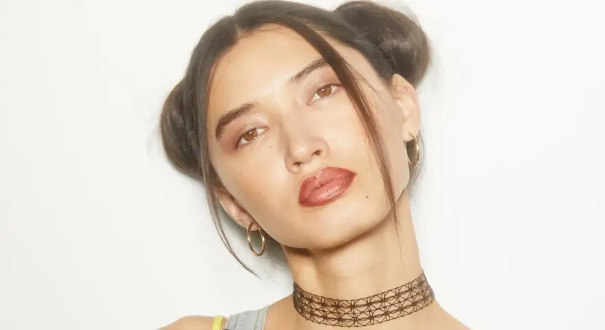 Chica con peinados y maquillaje de los 90, dos moños altos, labios marrones y choker.