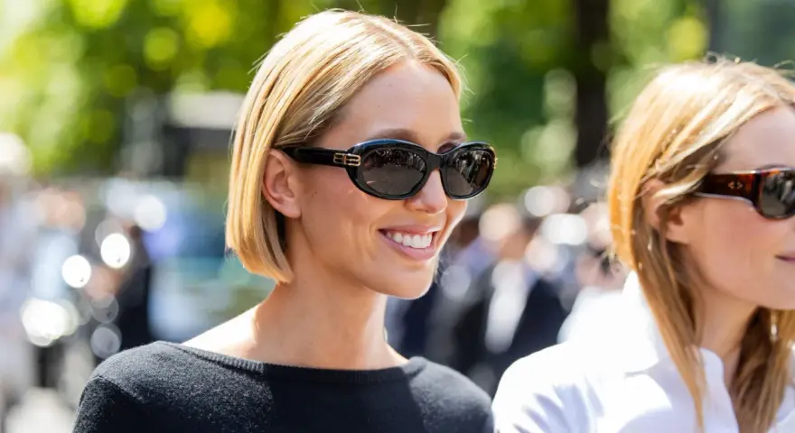 Mujer sonriente con cabello corto y cara redonda, luciendo gafas de sol negras y un suéter negro.