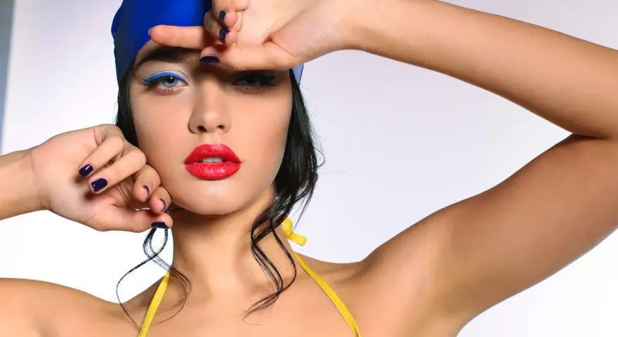 Mujer con gorro azul y traje de baño amarillo, labios rojos y delineado azul vibrante. Un look de maquillaje para ir a la playa.