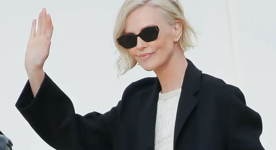 Mujer con cortes de pelo corto rubio, gafas de sol y chaqueta negra saluda.