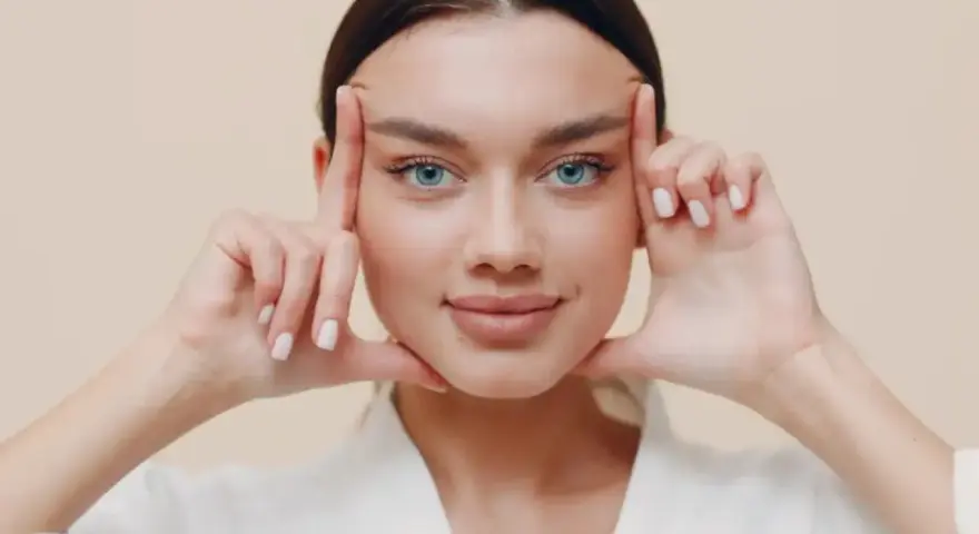Mujer con ojos azules realizando ejercicios faciales, aplicando presión en las sienes y barbilla para tonificar.