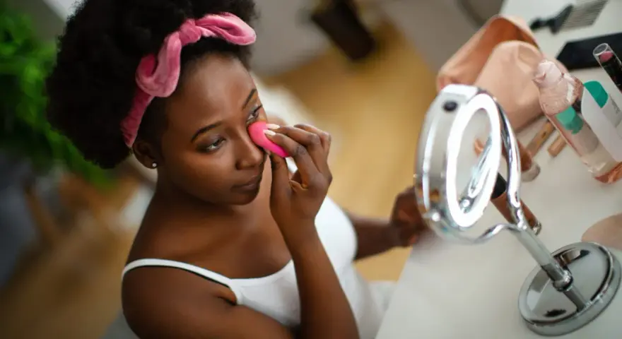 Una mujer se aplica base de maquillaje con esponja frente a un espejo.
