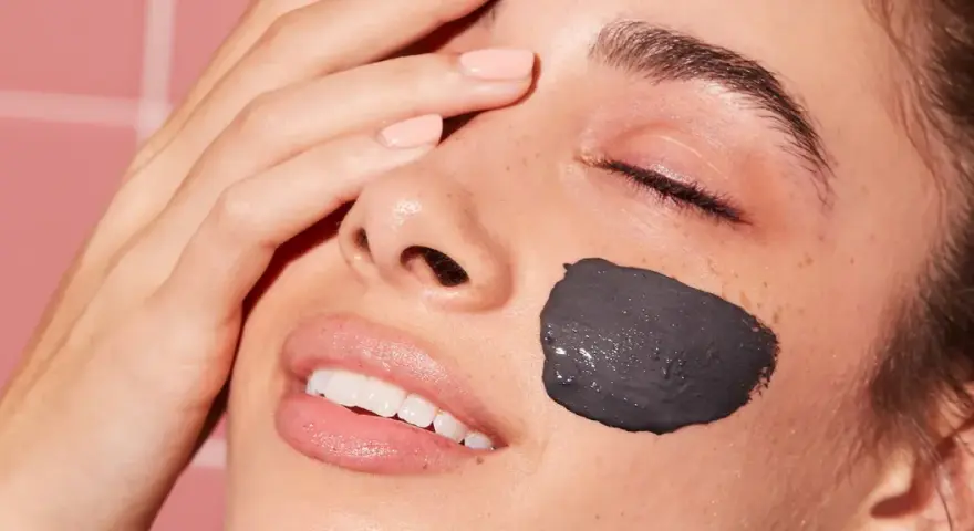 Una mujer sonríe con una mascarilla facial de arcilla negra en la mejilla. Después de una mascarilla facial, hay que ponerse crema para hidratar la piel.