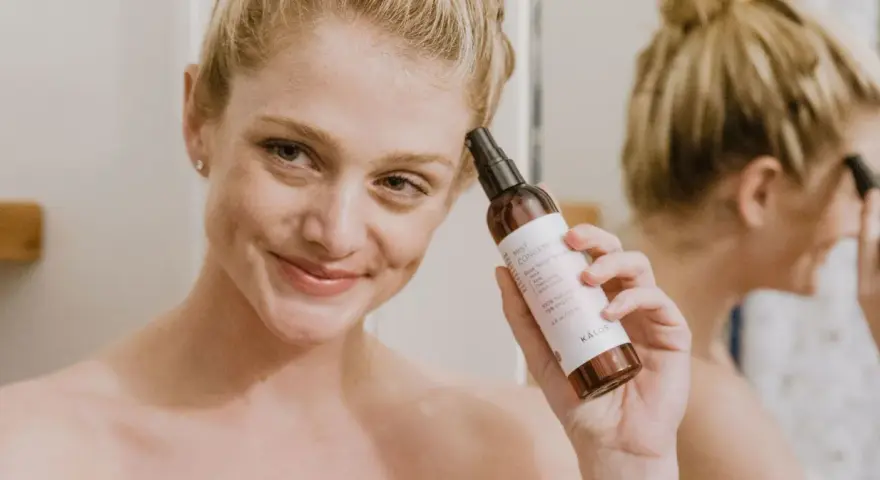 Mujer sonriente aplica bruma facial para como fijar el maquillaje. El producto es de la marca KÁLOS.