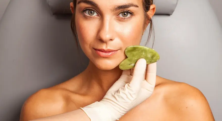 Una mano enguantada aplica una piedra de jade en la mejilla de una mujer, mostrando los beneficios del masaje facial para una piel radiante.