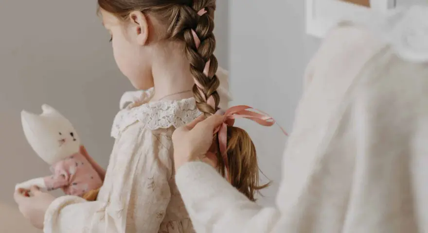 Manos terminan de trenzar el cabello de una niña, añadiendo cintas rosas. Ideas de peinados para niñas adorables.