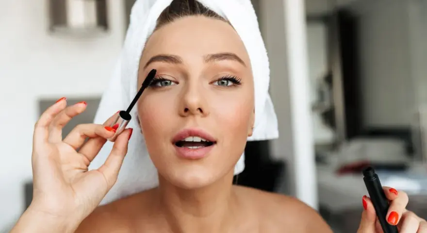 Mujer con toalla en la cabeza aplicándose máscara de pestañas como parte de su rutina maquillaje.
