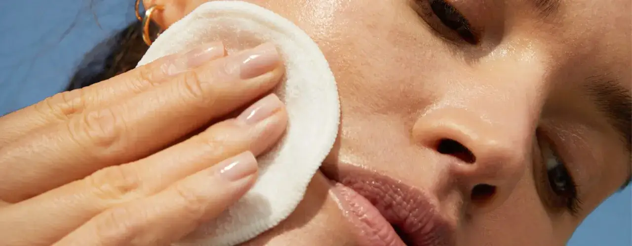 Una mano con uñas cuidadas usa un disco de algodón para retirar el maquillaje. El agua micelar se enjuaga suavemente, dejando la piel radiante.