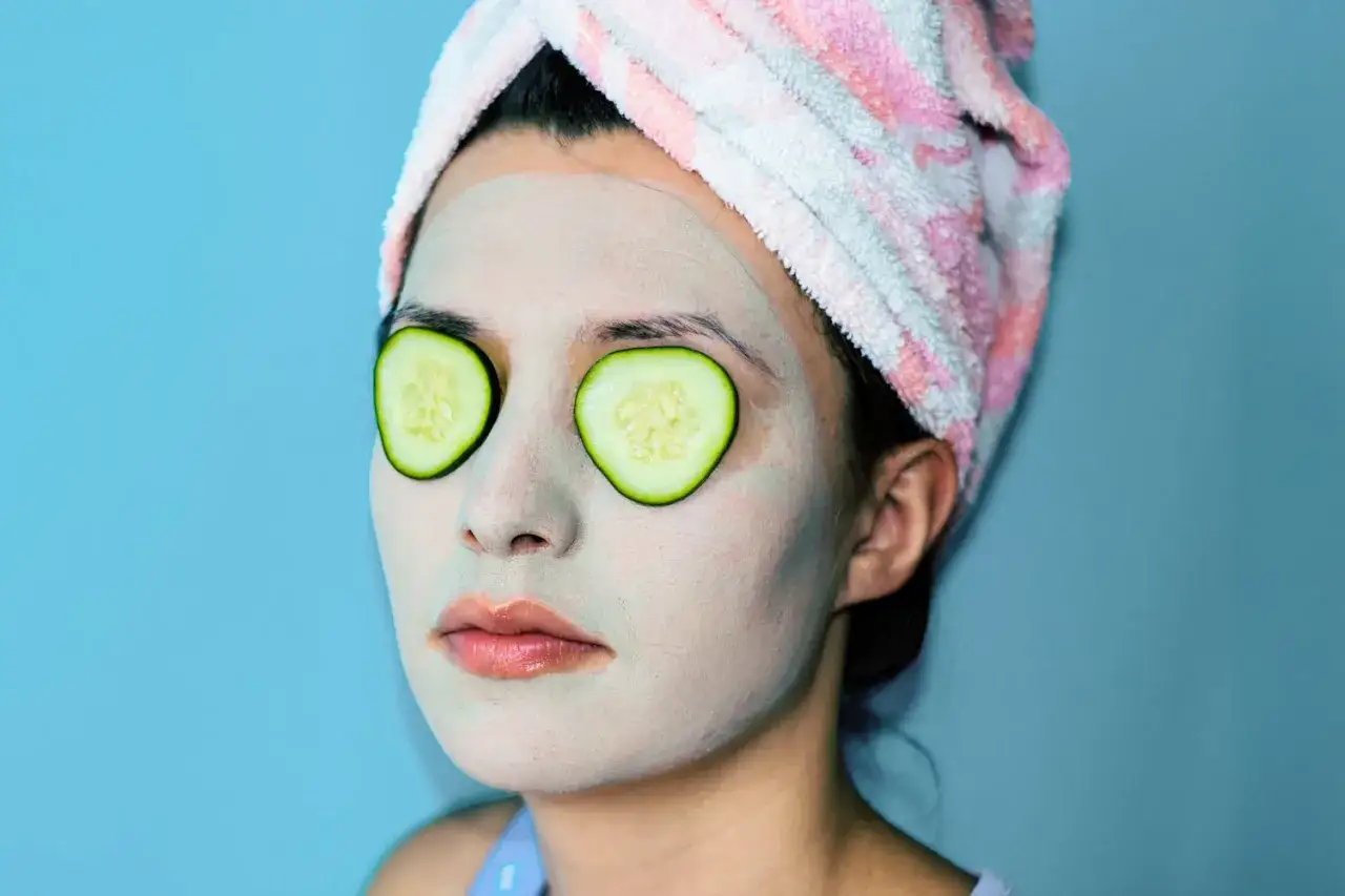 Mujer con mascarilla facial y rodajas de pepino en los ojos, envuelta en una toalla.