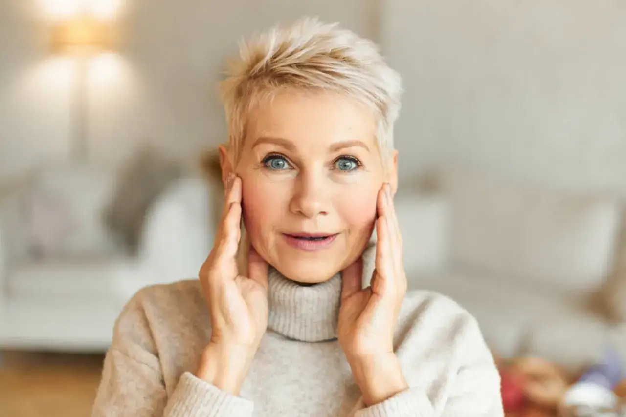 Mujer con cabello rubio corto y ojos azules, aplicando maquillaje natural para piel madura.