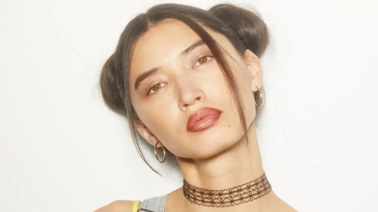 Chica con peinados y maquillaje de los 90, dos moños altos, labios marrones y choker.