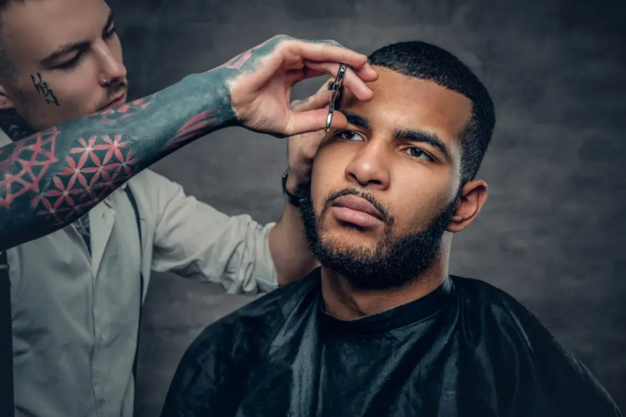 Barbero con tatuajes en el brazo recorta el buzz cut corto de un hombre con barba.