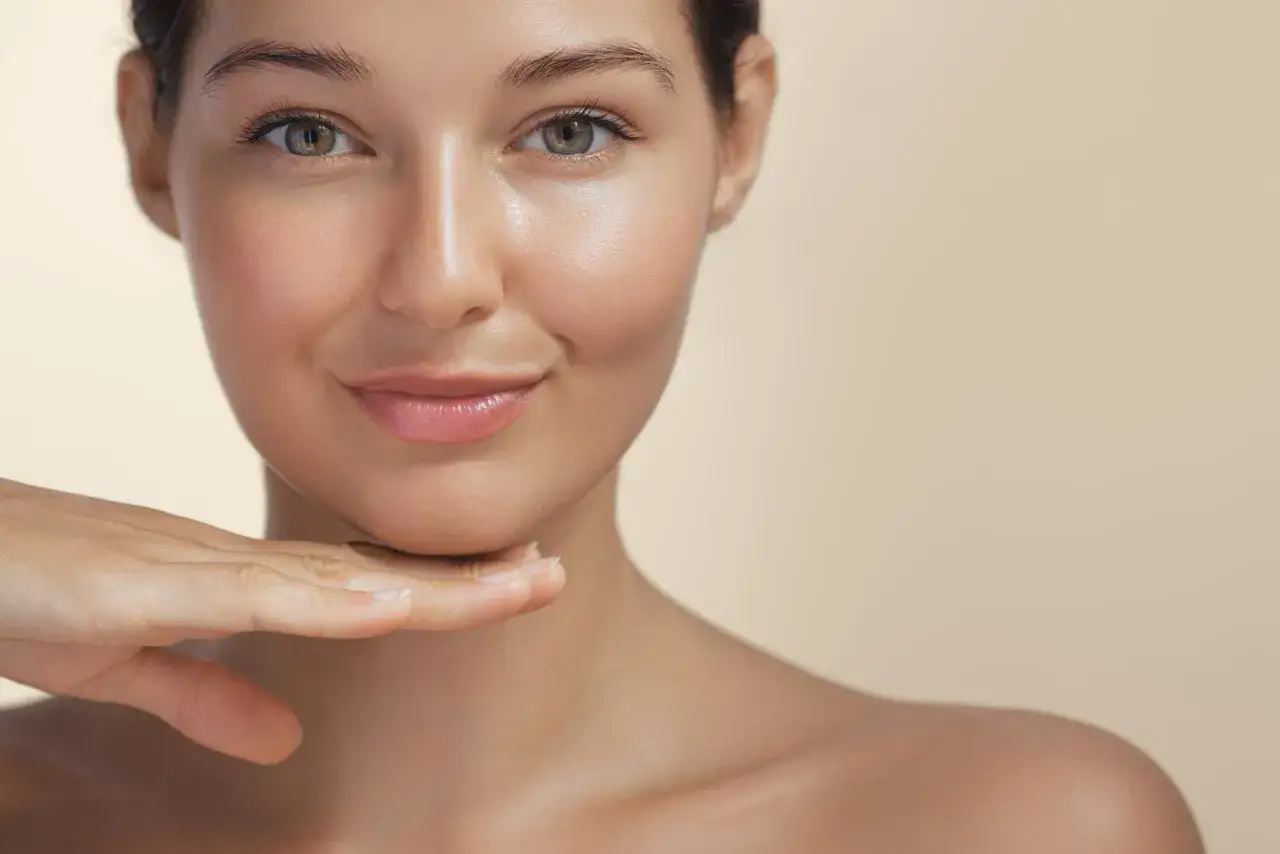 Rostro de mujer con piel radiante, un toque de mano en la barbilla. Su piel refleja la luz, mostrando un glow natural y saludable.