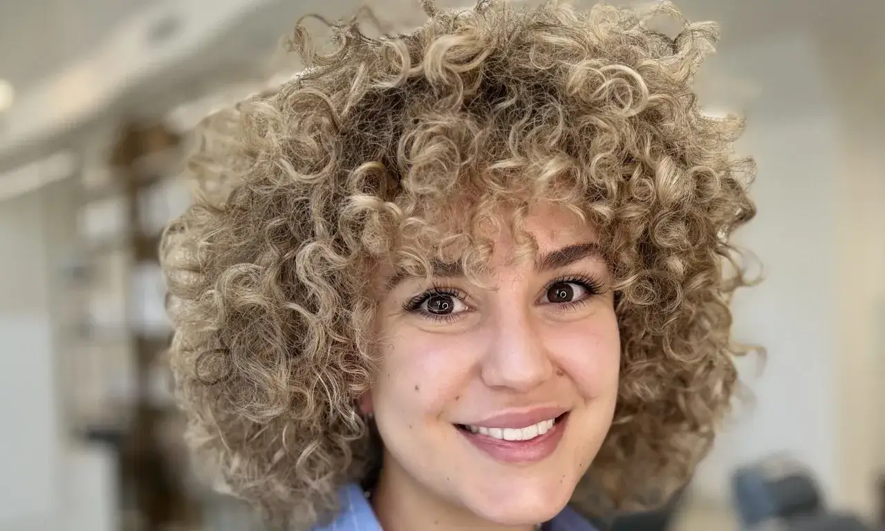 Mujer sonriente con cabello rizado rubio y voluminoso. Un ejemplo de los diversos tipos de flequillo para pelo rizado que realzan la textura natural.