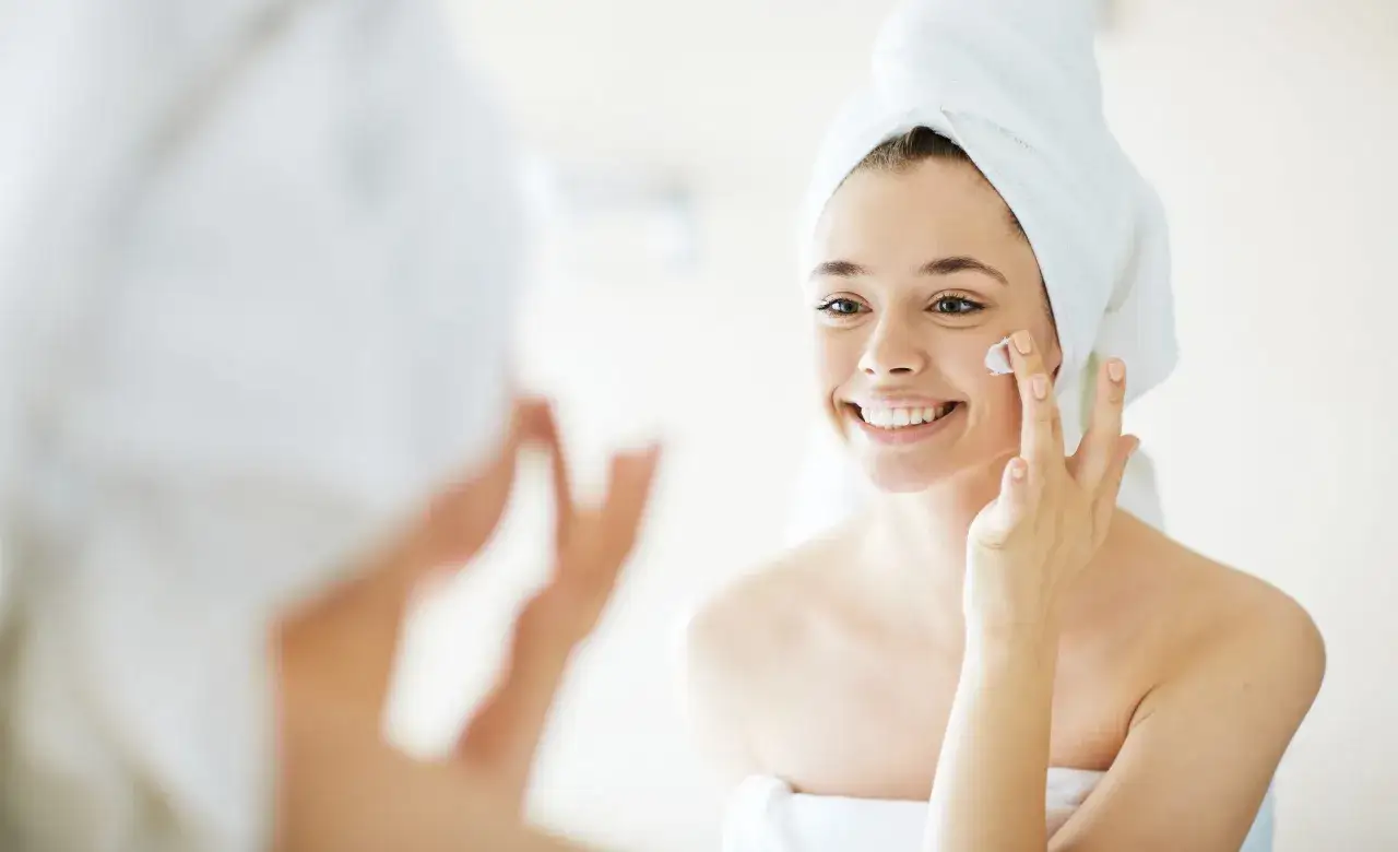 Mujer sonriente con toalla en la cabeza, aplicándose crema en la mejilla como parte de su rutina de limpieza facial.