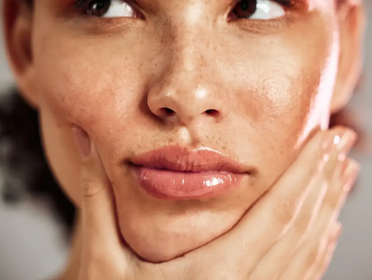 Primer plano de un rostro con labios brillantes y pecas en la nariz, con una mano acariciando la mejilla.