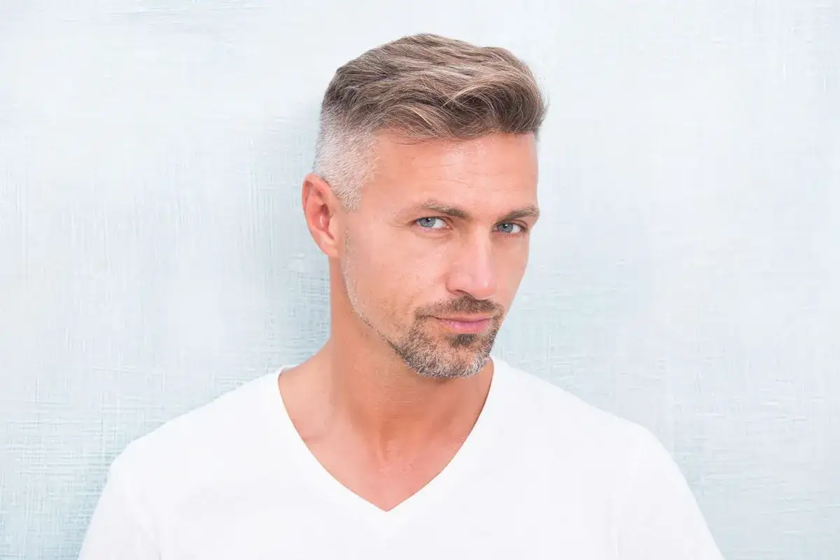 Hombre con perilla barba y cabello peinado, viste camiseta blanca.