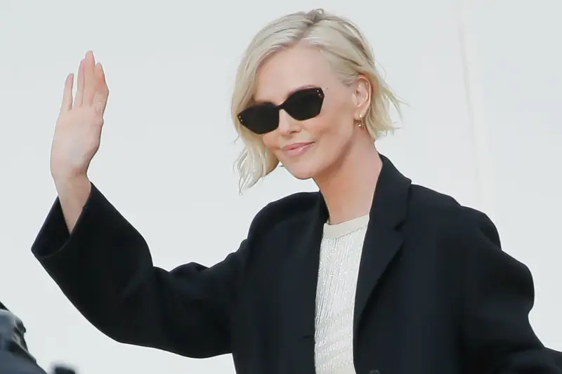Mujer con cortes de pelo corto rubio, gafas de sol y chaqueta negra saluda.
