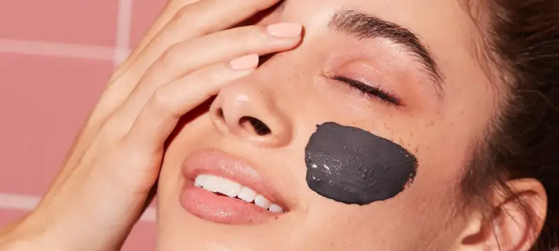 Una mujer sonríe con una mascarilla facial de arcilla negra en la mejilla. Después de una mascarilla facial, hay que ponerse crema para hidratar la piel.